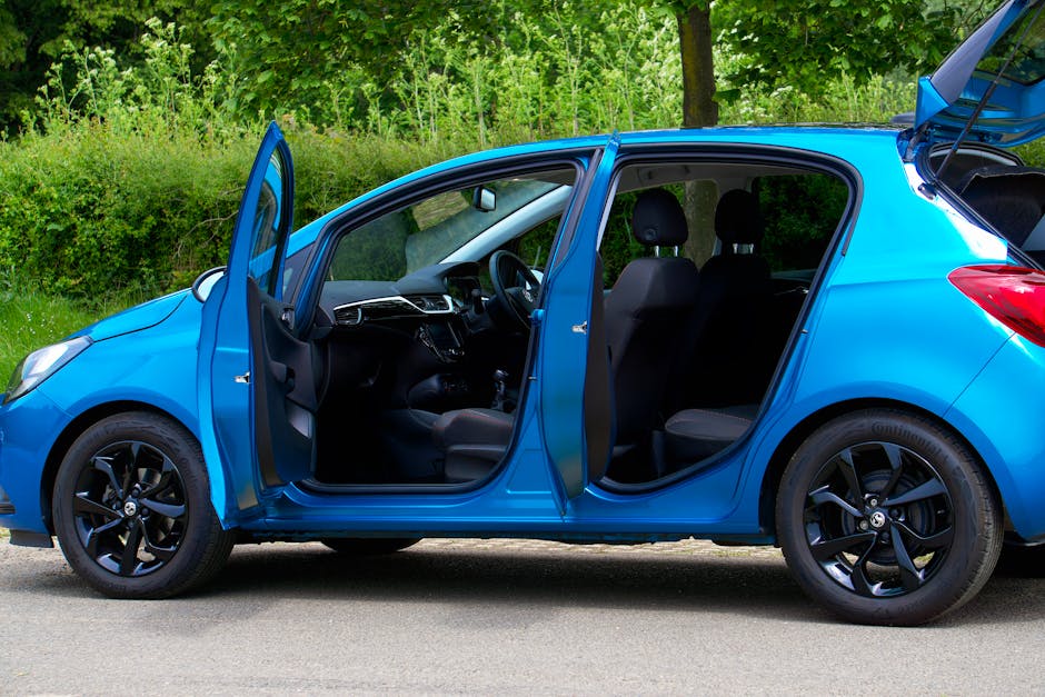 A blue compact hatchback car parked on a paved road outdoors, with its front left and rear passenger doors open, revealing a dark interior with fabric seats and a steering wheel. The vehicle's black a