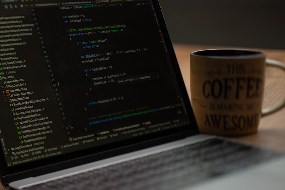 A close-up view of a laptop screen displaying lines of code in various colors, positioned on a wooden desk. Next to the laptop, there is a ceramic coffee mug with a beige and brown color scheme, featu