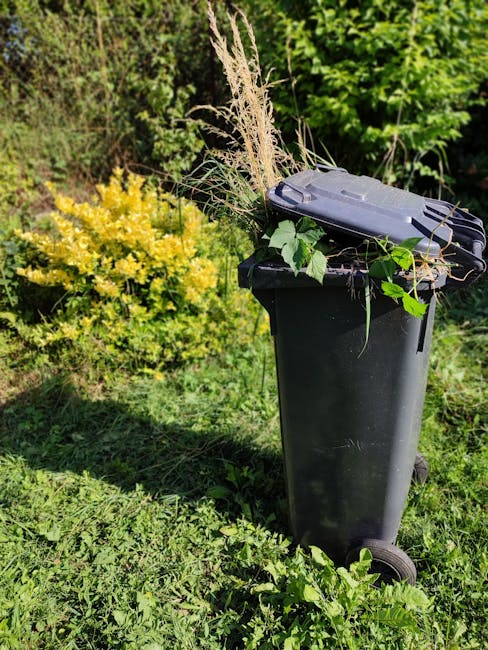 A black wheeled garden waste bin standing on a grassy outdoor area, filled with cut grass, dry ornamental grasses, and leafy plant stems. Surrounding the bin are vibrant green shrubs and a small bush 